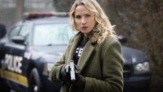 Nina stands by a squad car with gun drawn and an FBI vest under her green coat