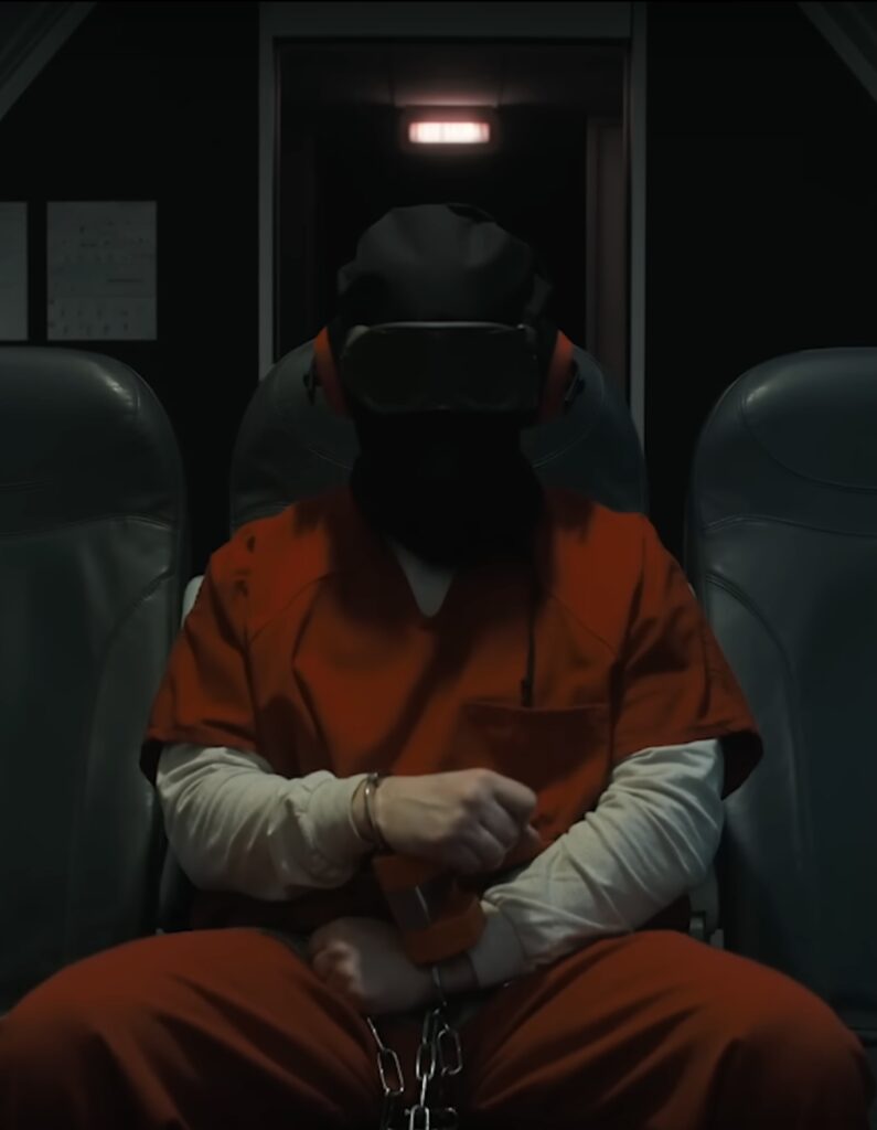 A hooded prisoner in an orange jumpsuit shackled and wearing headphones