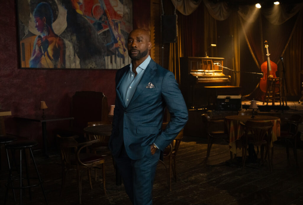 Watson in piedi nel club con le mani in tasca in Watson Stagione 2 Episodio 7