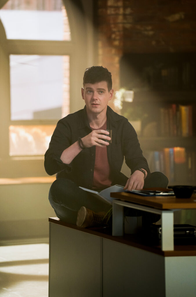 Adams sitting at a desk drinking something on Watson Season 2 Episode 8